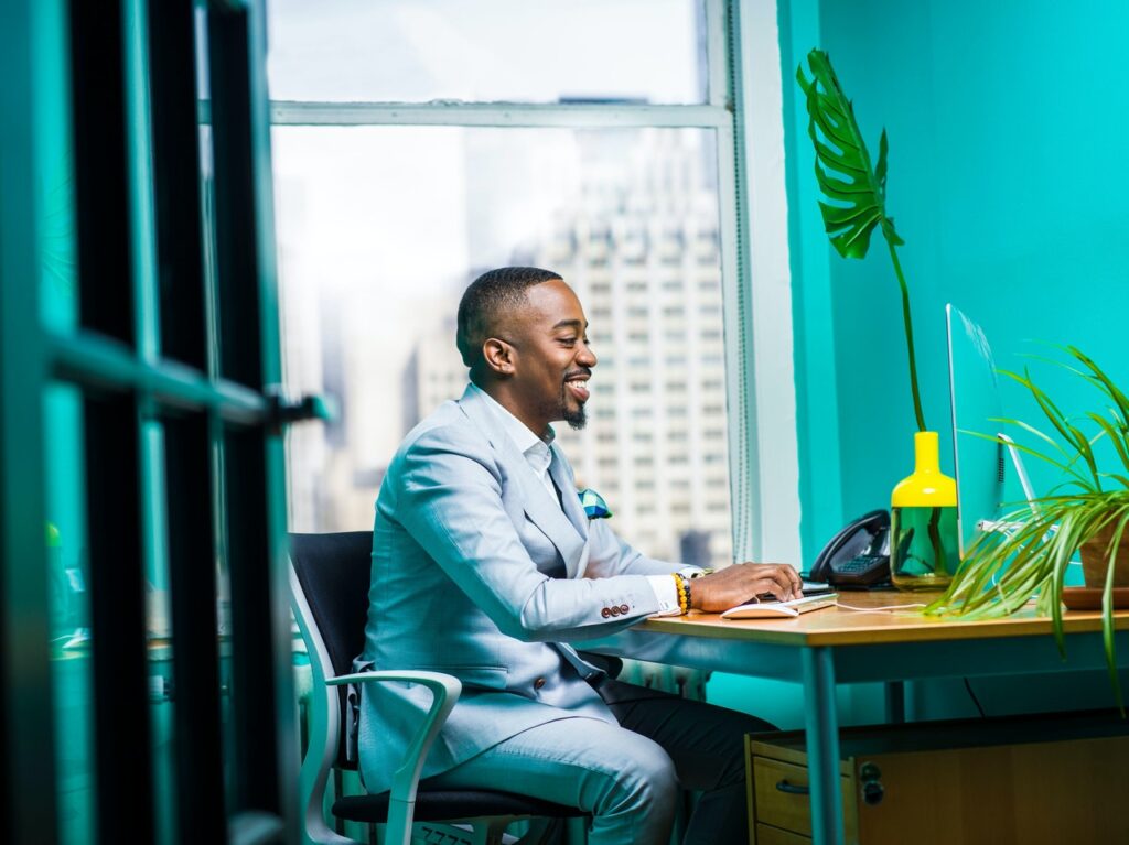 Um homem negro, vestindo terno cinza, está sentado em um escritório, mexendo em um computador. Ele sorri como se estivesse relaxado. É possível ver algumas plantas no ambiente.