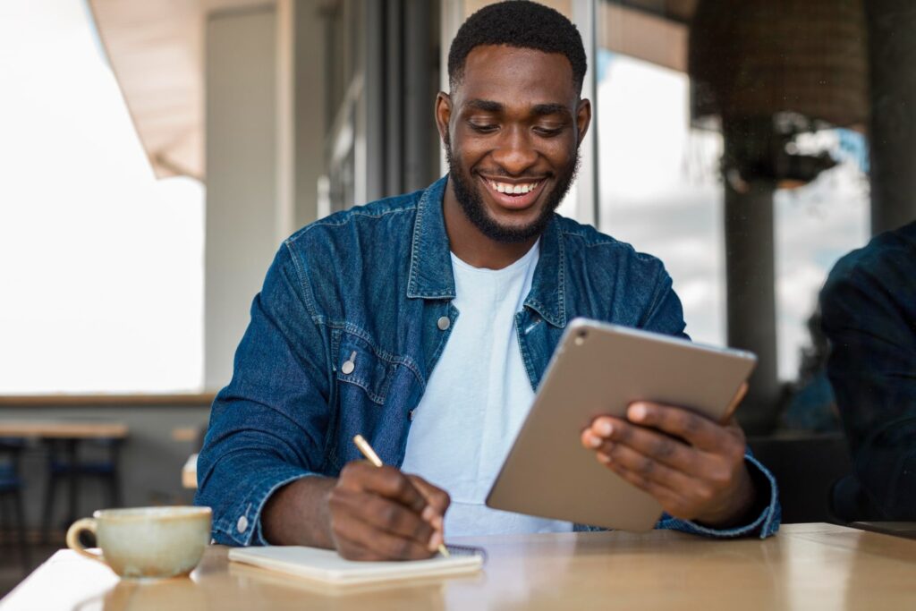 Um homem negro está sentado em uma mesa de restaurante, enquanto mexe em um tablet com a mão esquerda e faz anotações em uma agenda com a mão direita. Ele está sorrindo. Há uma xícara de café sobre a mesa.