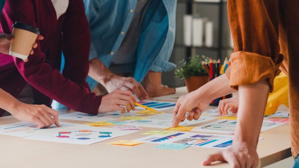Um grupo de pessoas está em um escritório conversando. As pessoas olham e apontam para várias folhas de papel com gráficos de desempenho sobre uma mesa.