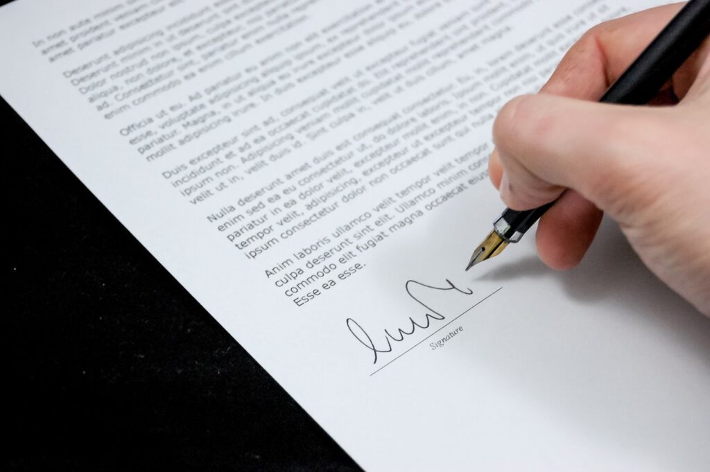 É possível ver a mão de uma pessoa segurando uma caneta e fazendo uma assinatura em um contrato.