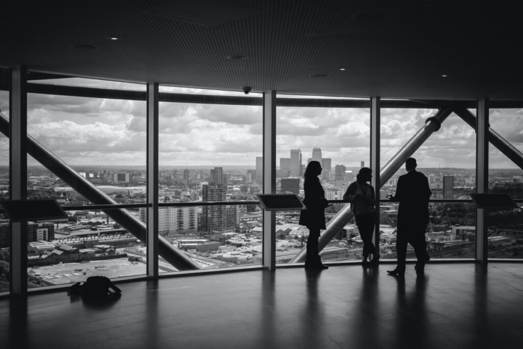 Três pessoas conversam e olham pela fachada de vidro de uma sala de escritórios vazia de um prédio. A imagem está em preto e branco. 