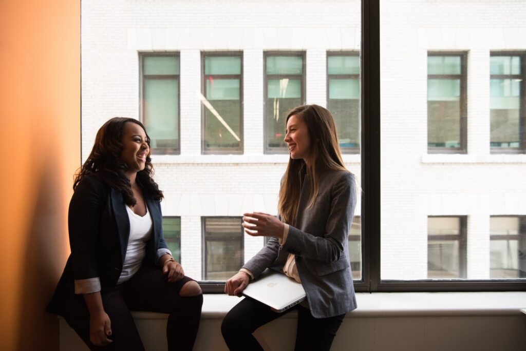 Duas mulheres, sendo uma branca e uma negra, estão sentadas no beiral da janela de um prédio. Elas conversam e riem. A mulher branca carrega um laptop.
