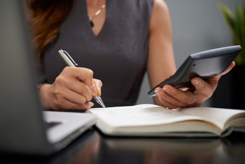 Uma mulher está sentada em uma mesa de escritório, usando uma calculadora na mão esquerda e uma caneta na mão direita. Ela faz anotações em uma agenda. Há um laptop prateado à sua frente.