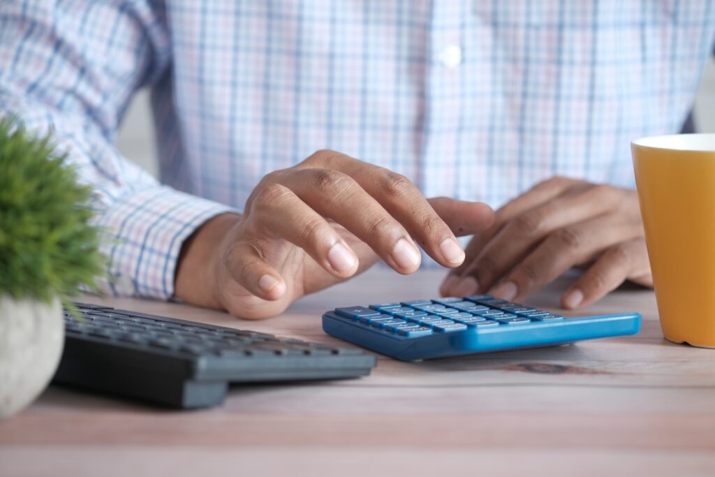 Uma pessoa negra, vestindo camiseta social branca com estampa quadriculada azul-claro, está mexendo em uma calculadora também azul. Ao seu lado, um teclado de computador na cor preta, um vaso com planta e um copo de café.