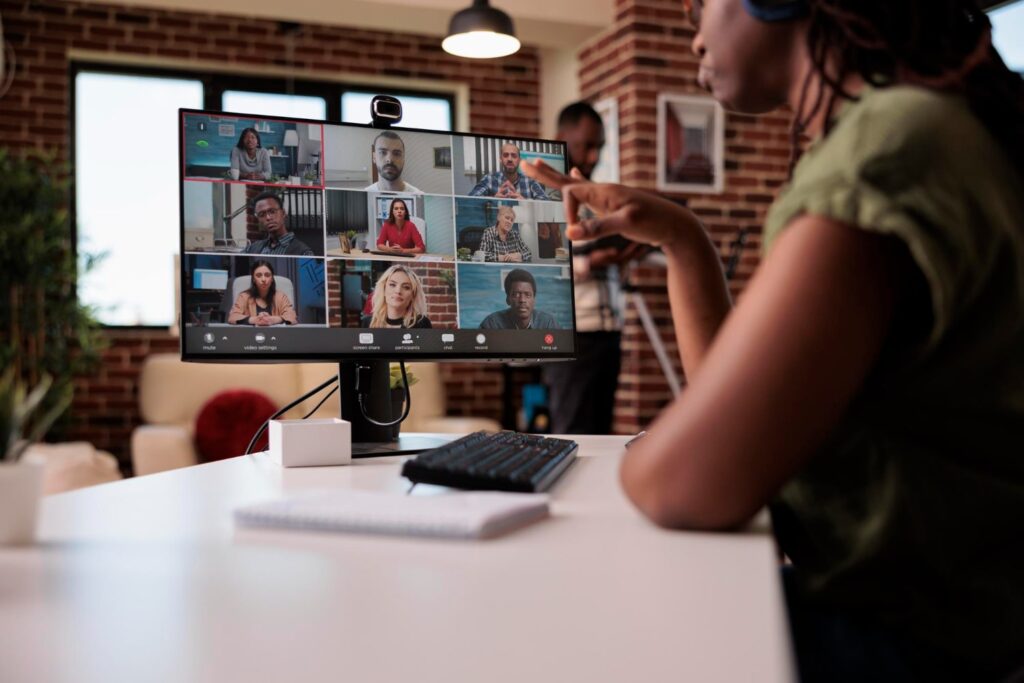 Uma mulher negra, sentada em uma mesa de escritório, tendo uma reunião remota pelo computador. Na tela do aparelho, é possível ver outras nove pessoas. Ao fundo, fora de foco, há um homem, também negro, em pé.
