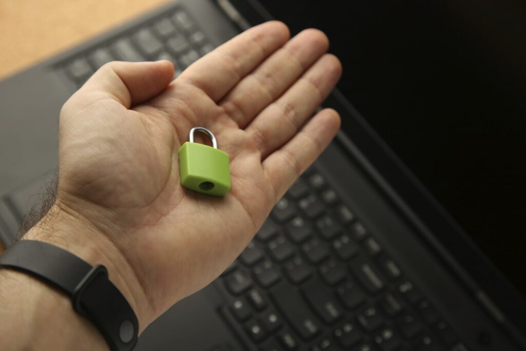 É possível a mão de uma pessoa branca, segurando um cadeado verde-claro. Essa pessoa usa um relógio de pulso e está com a mão suspensa sobre um laptop preto.
