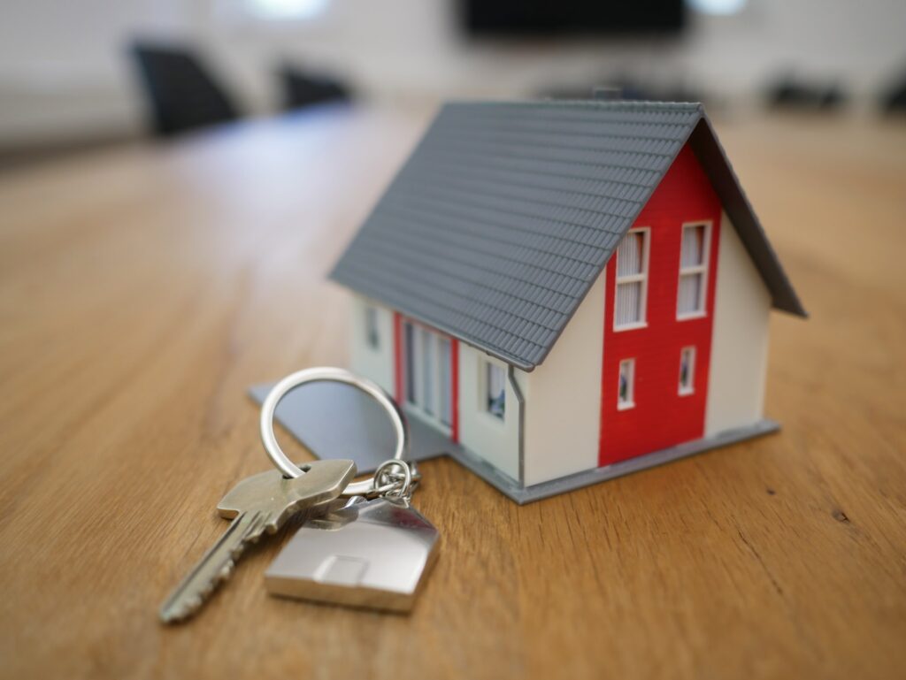 É possível ver uma chave de porta e um chaveiro em formato de casa sobre uma mesa de madeira. Ao lado, uma casinha em miniatura. Ao fundo, fora de foco, o que parece ser um escritório ou sala de reuniões.