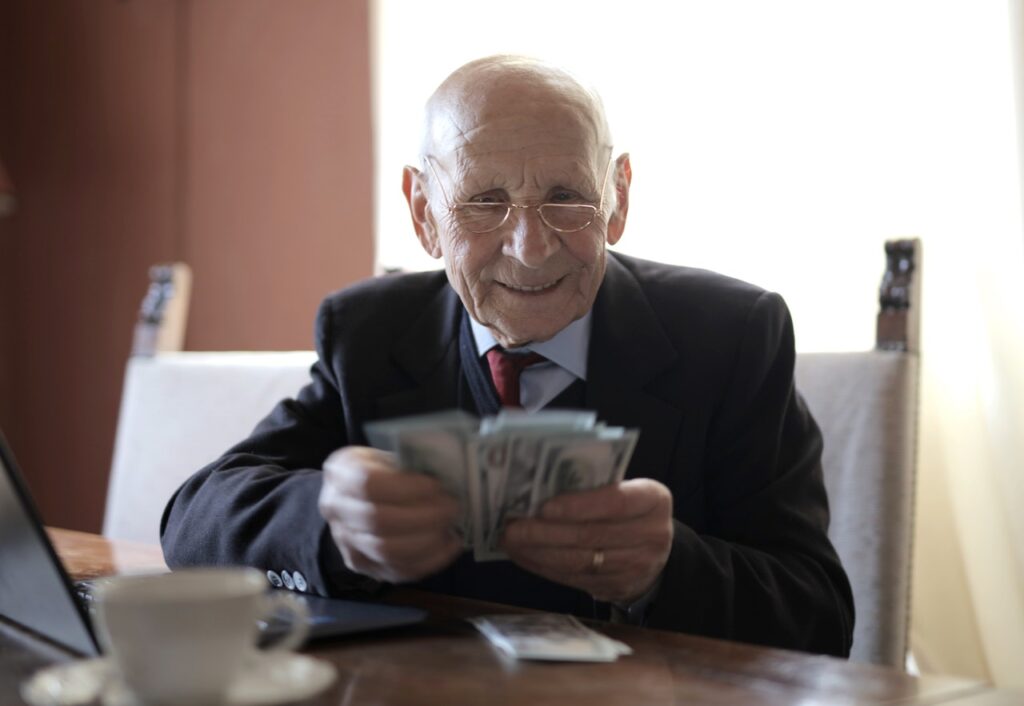 Um homem branco, já idoso, vestindo terno e usando óculos, está sentado em um escritório, contando várias notas de dinheiro. À frente dele, há uma mesa com um laptop, uma xícara de café e mais algumas notas.